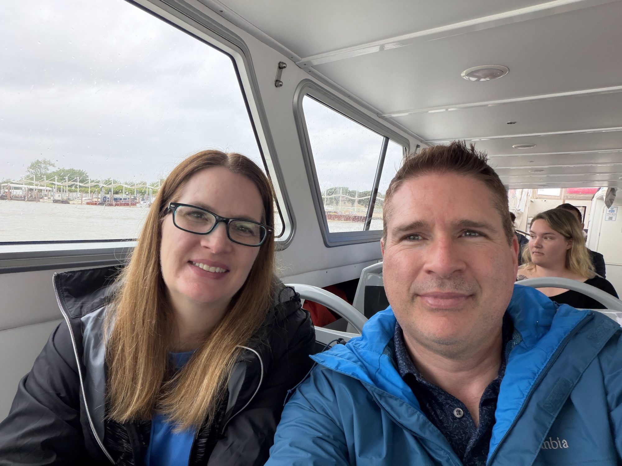 Close selfie of Kyle and Mandy seated inside a ferry, rain jackets on, lagoon light through the windows