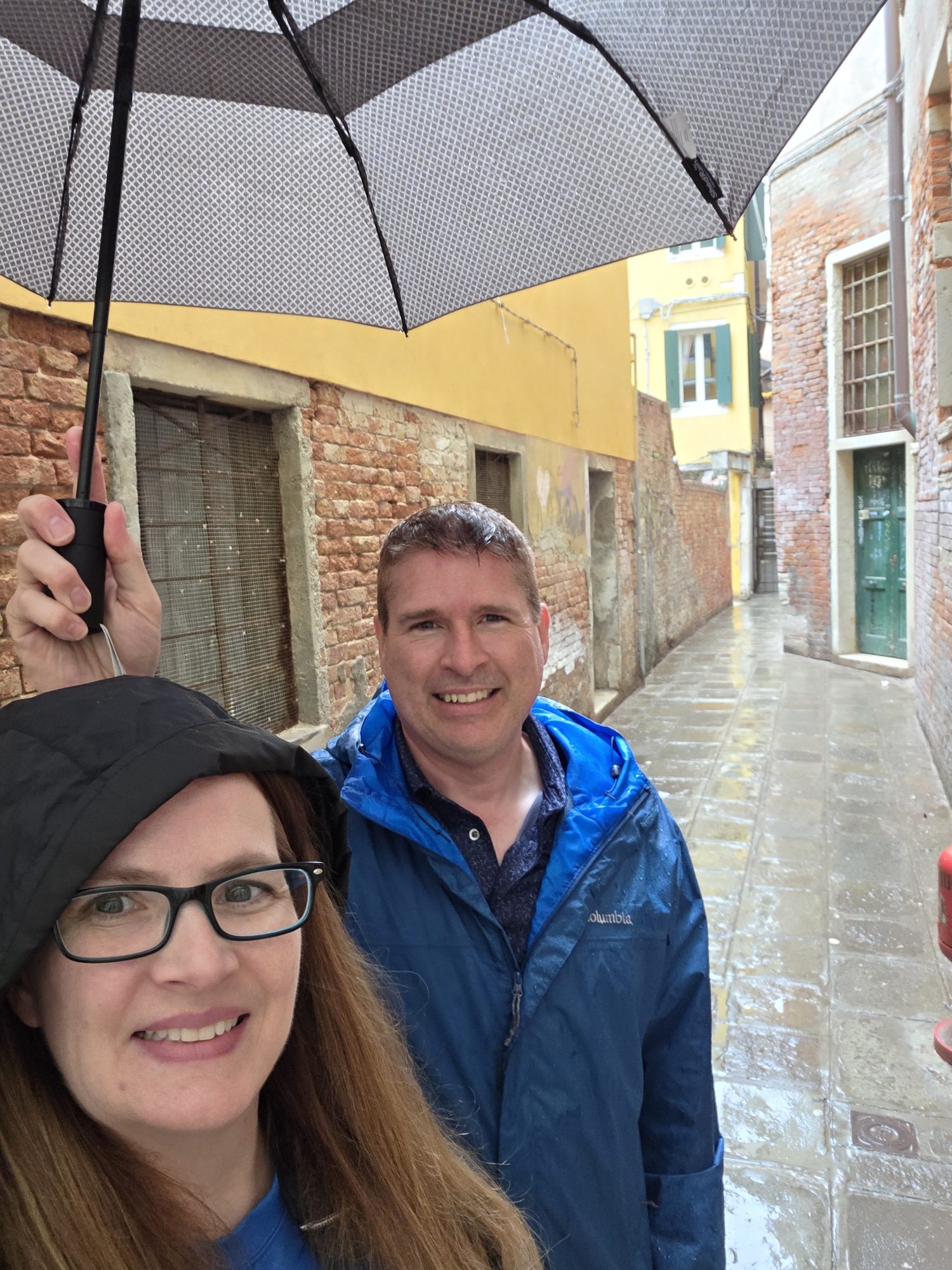 Kyle and Mandy under a checkered umbrella in a narrow rainy Venetian alley of brick and plaster