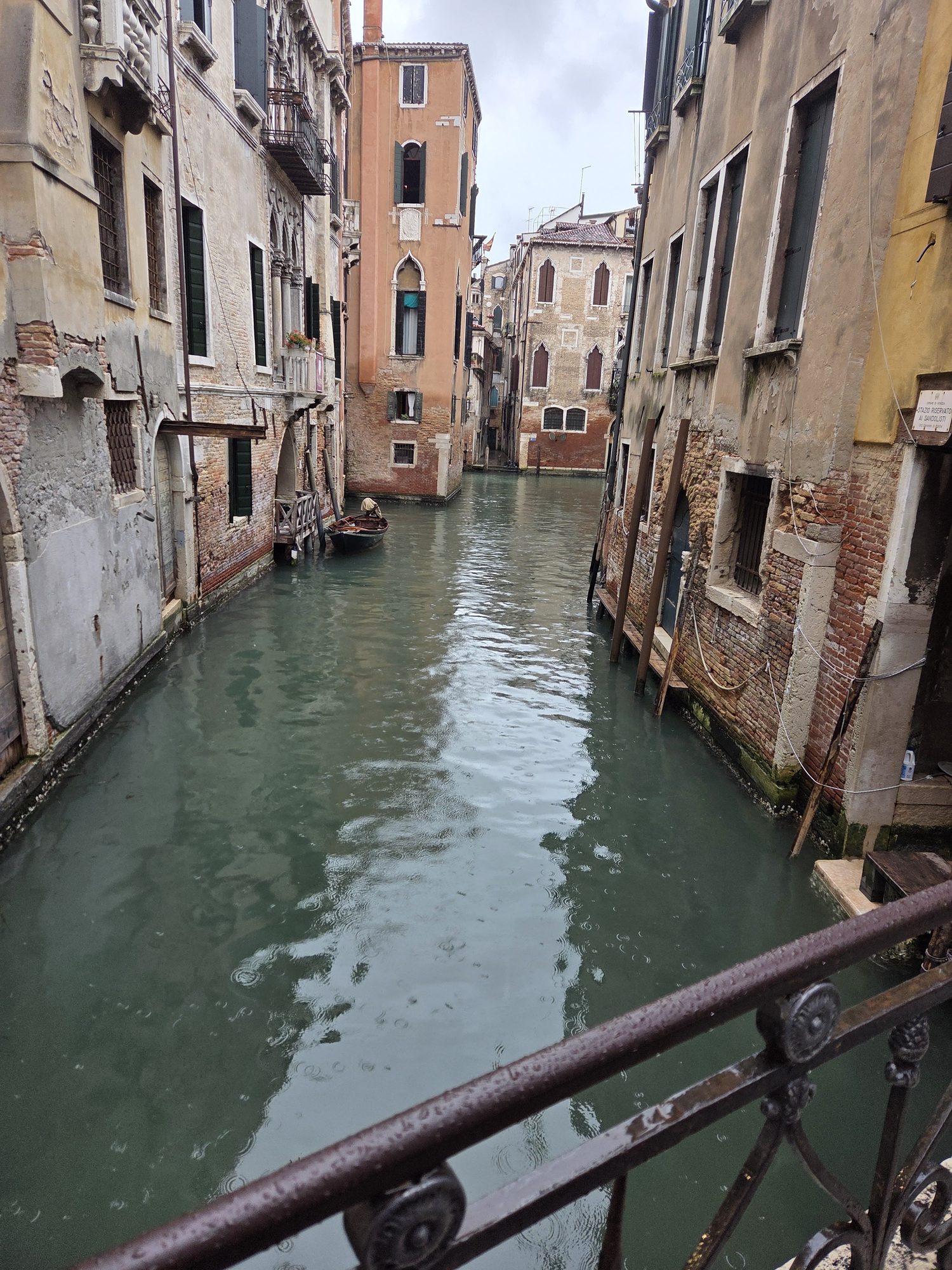 Narrow Venetian canal viewed over a wrought-iron bridge railing on a rainy overcast day