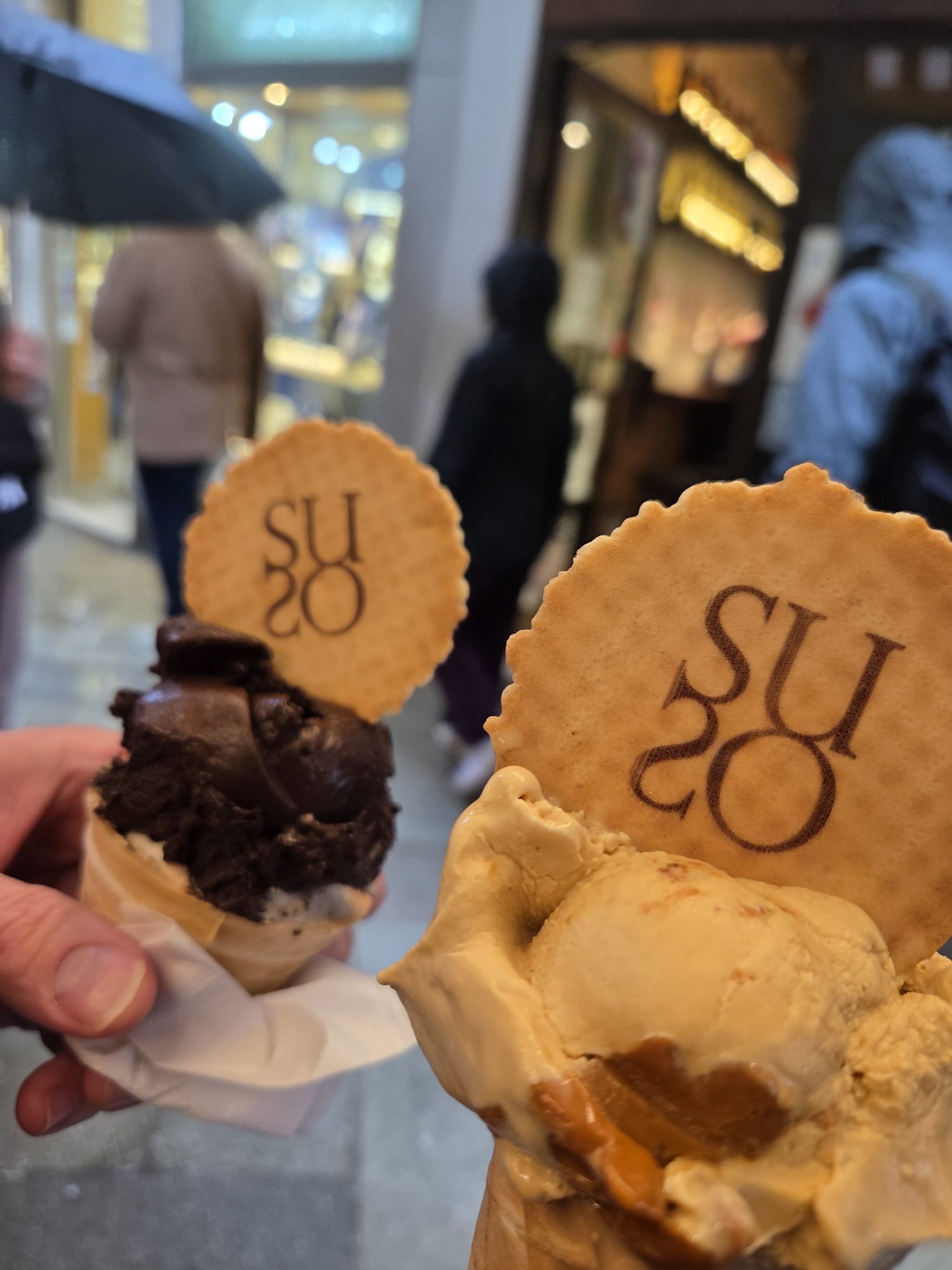 Two Suso gelato cones held up in a rainy Venice street, chocolate and a lighter flavor with Suso wafers