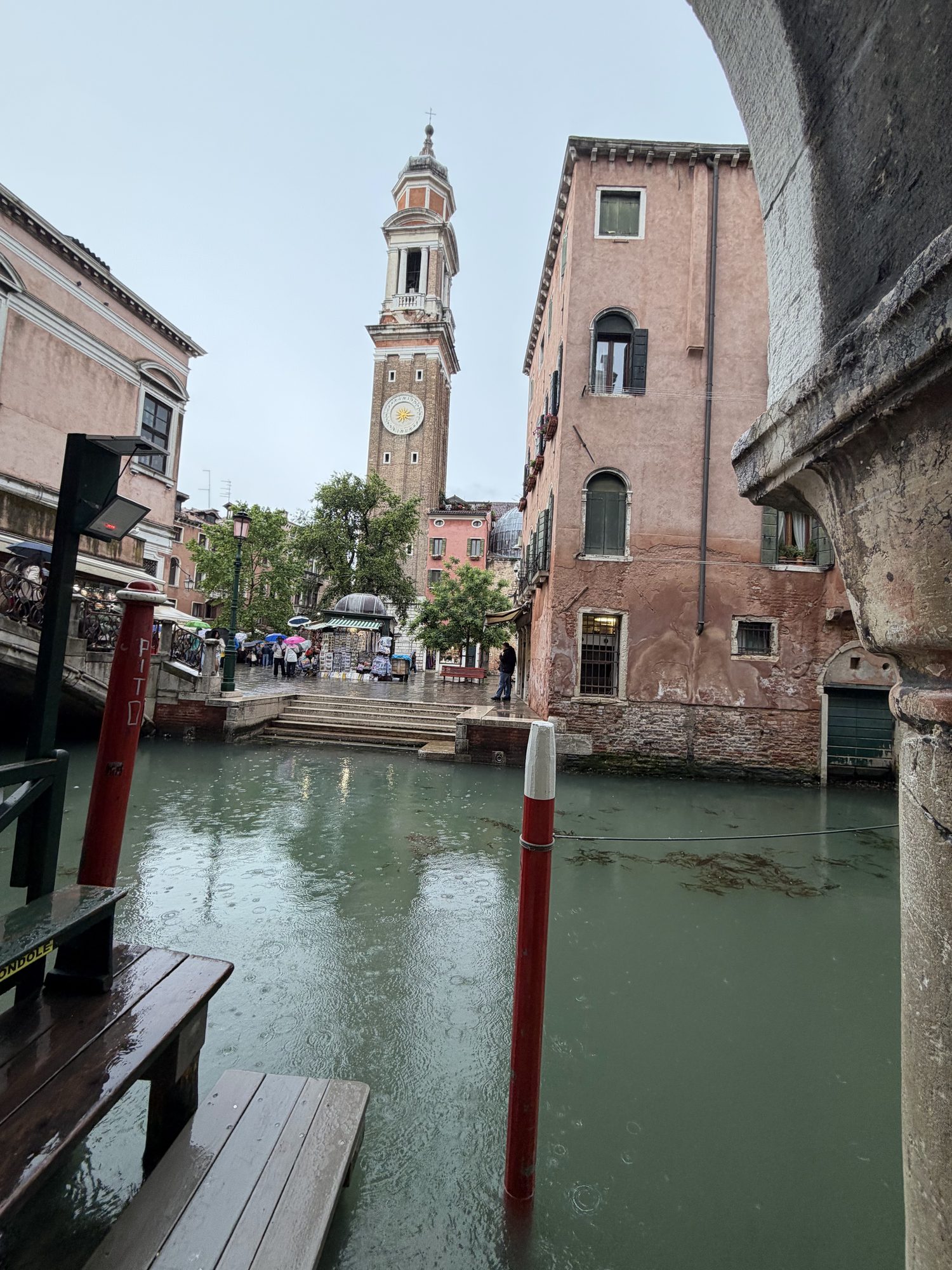 Rain ripples on a Venice canal with red-and-white mooring poles and the San Cassiano campanile beyond