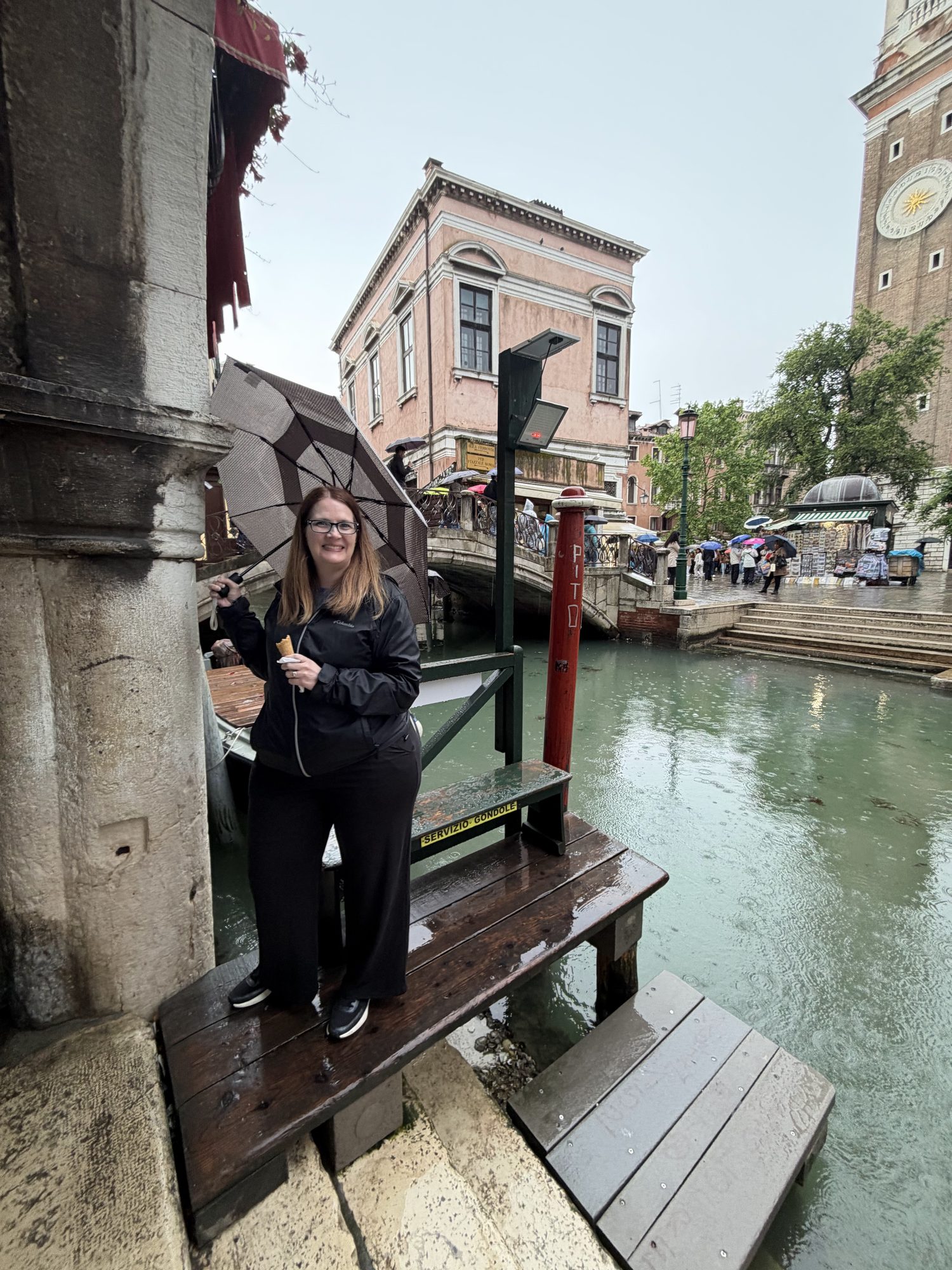 Mandy smiling on a wooden canal pier in Venice, holding gelato and a checkered umbrella in the rain