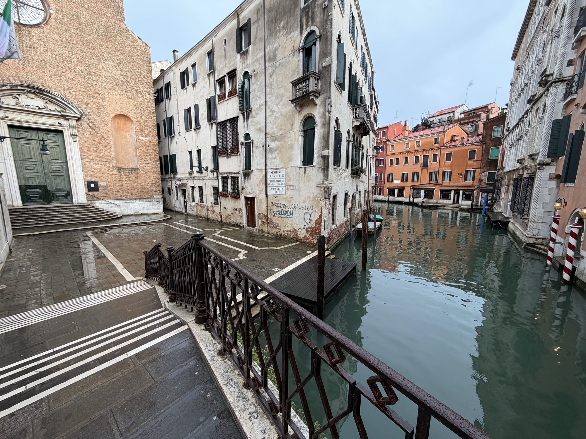 Quiet wet campo and canal beside a brick church entrance with green doors and striped mooring poles
