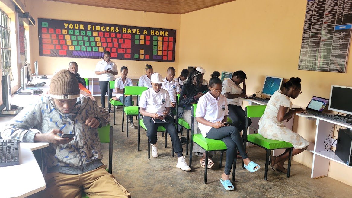 Students and staff working on laptops and phones in the institute computer lab beneath a typing skills poster