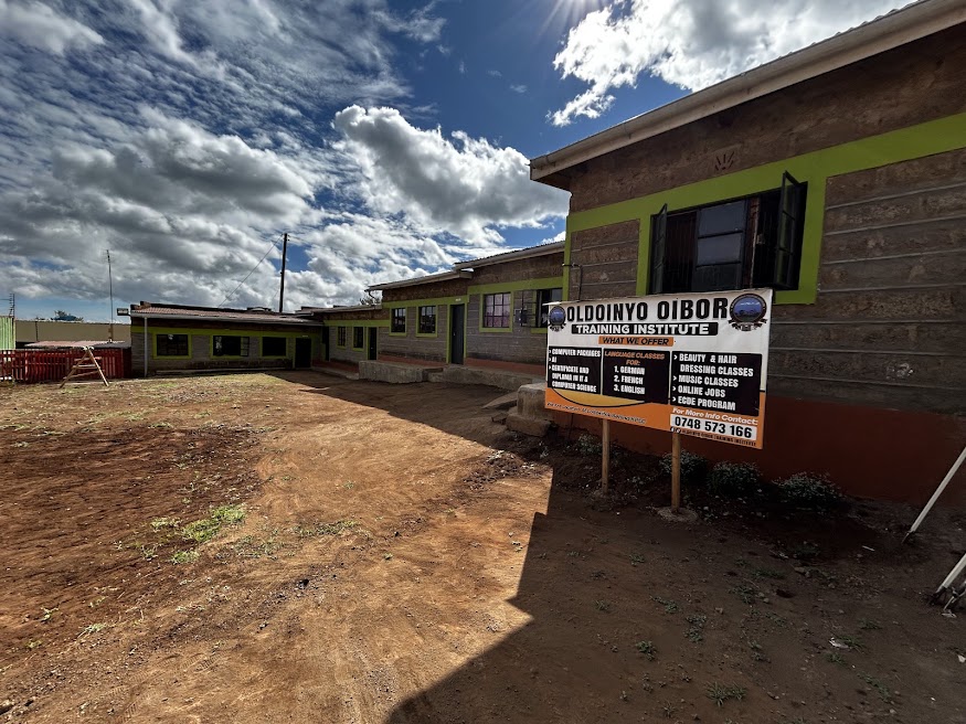 Exterior of Oldoinyo Oibor Training Institute with a courtyard sign listing computer, language, beauty, music, and ECDE programs
